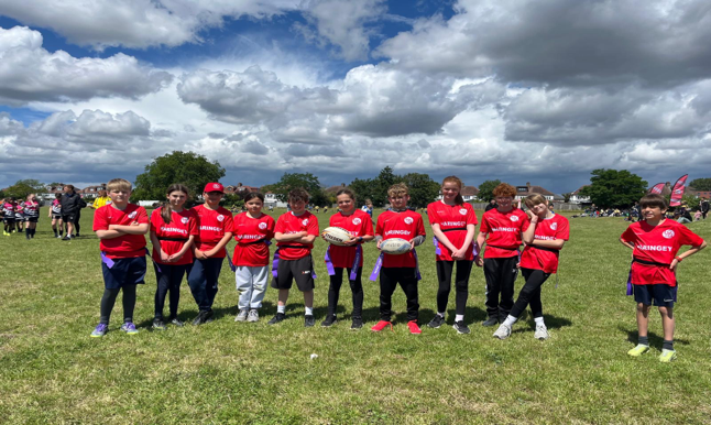 Group of children in a line wearing red t-shirts with purple hanging straps holding a rugby ball (tag rugby)